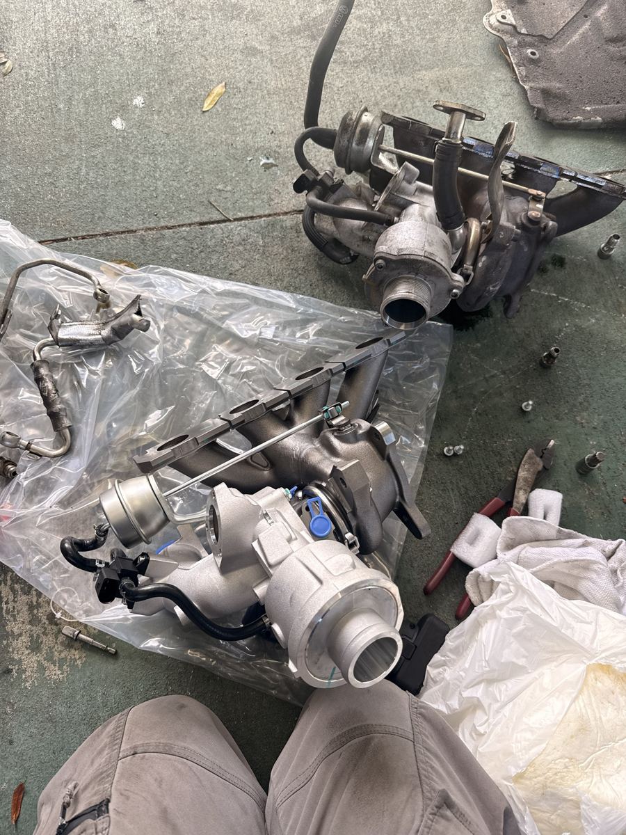 Old turbocharger next to a brand-new replacement turbo and manifold, laid out on a driveway